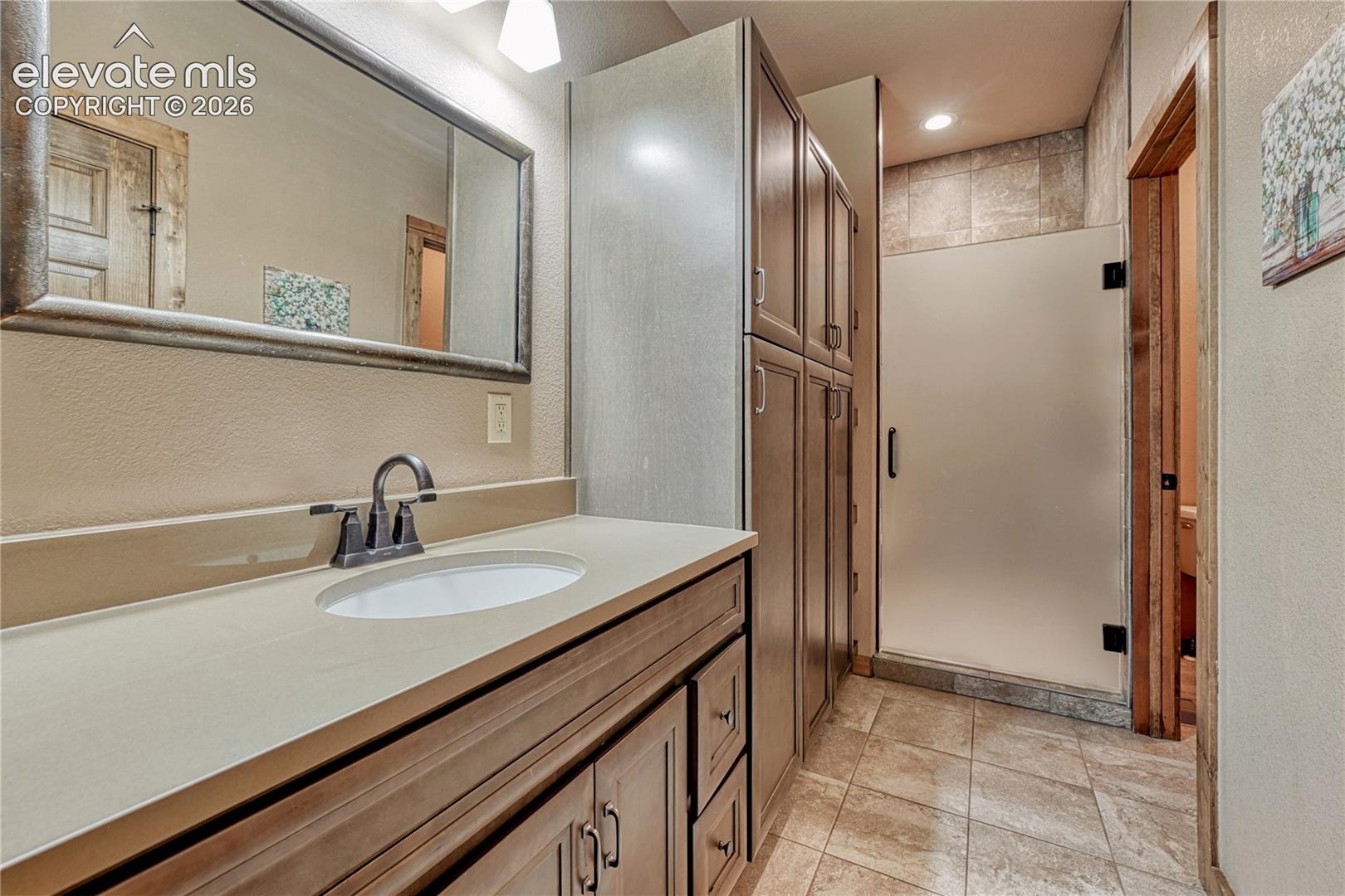 Bathroom featuring wood-finish cabinetry, a solid-surface countertop with an integrated basin, a full-height storage tower, and a walk-in shower with a frosted glass door