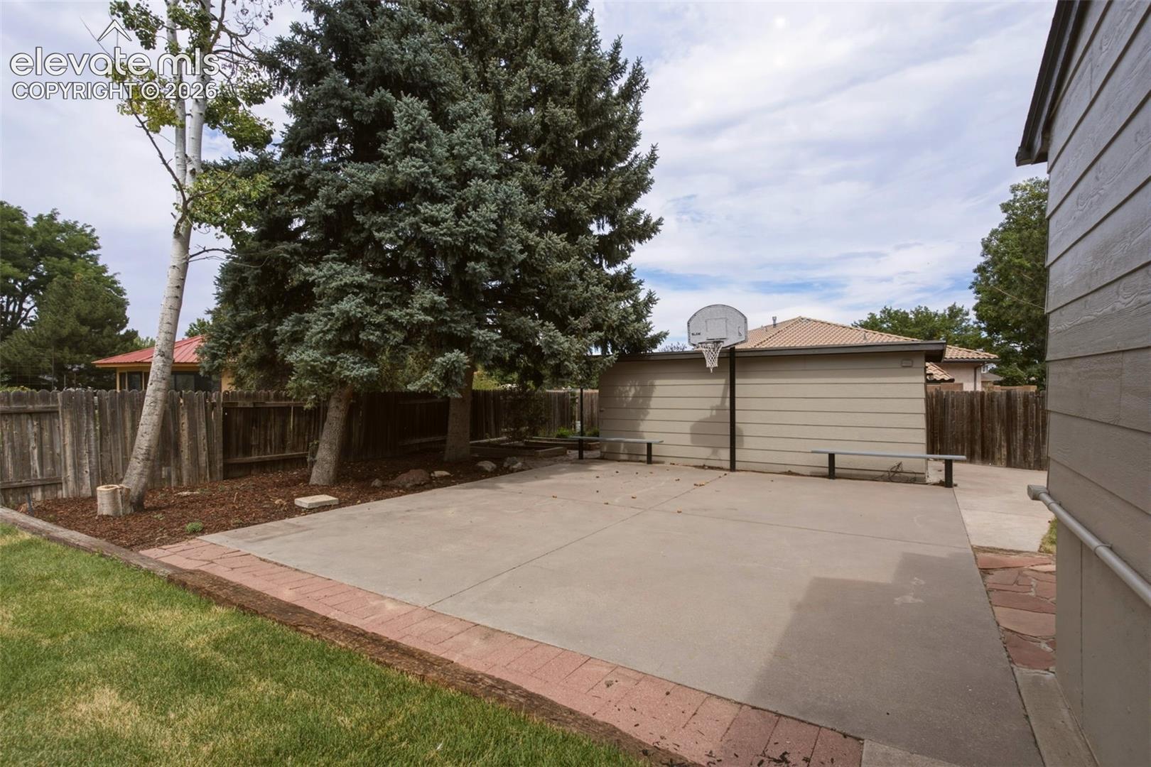 Paved patio area featuring a basketball hoop, mature trees, and a privacy fence
