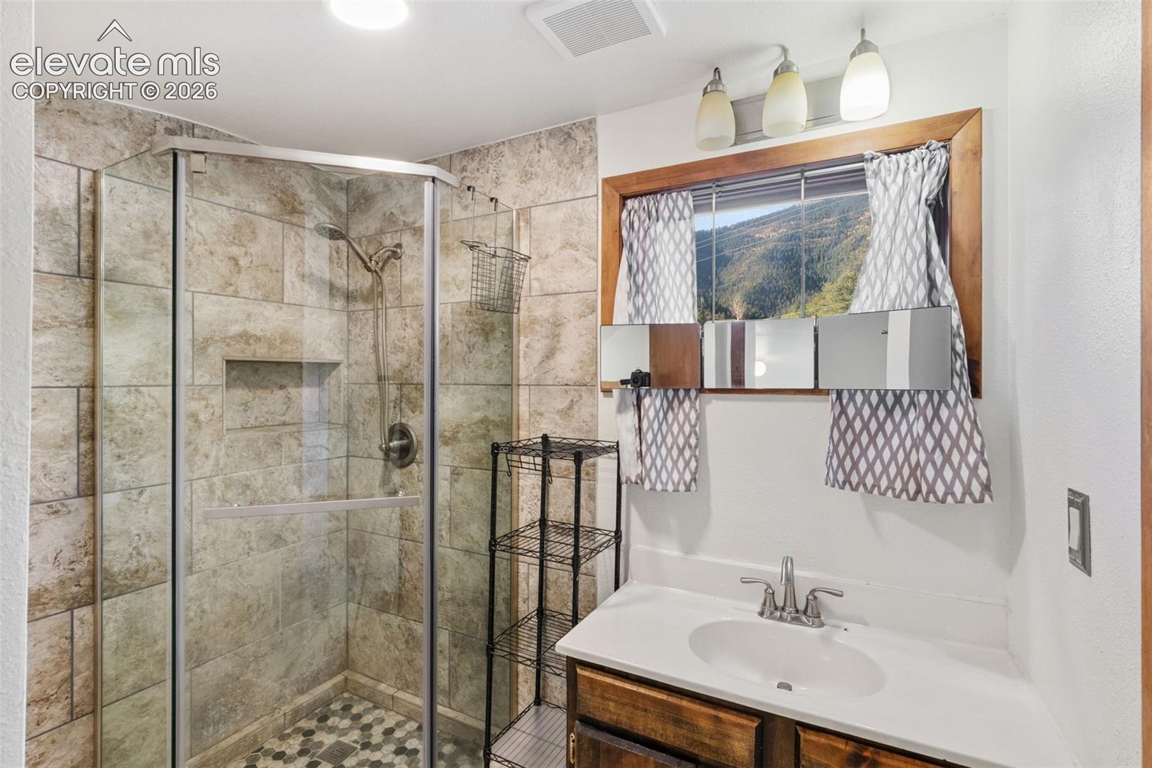 Corner shower stall with stone-finish tile surround and recessed shelving, wood-framed window featuring a mountain backdrop, white countertop with integrated oval sink, and three-light overhead vanity fixture