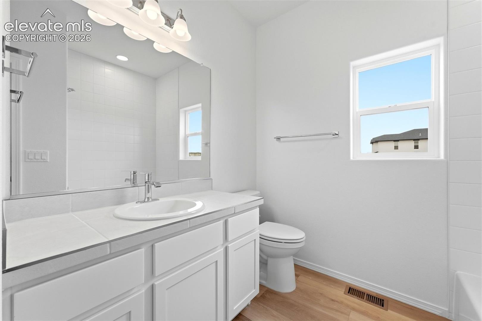 Bathroom featuring white shaker-style cabinetry, an integrated sink with a polished chrome faucet, a large vanity mirror, and wood-finish flooring