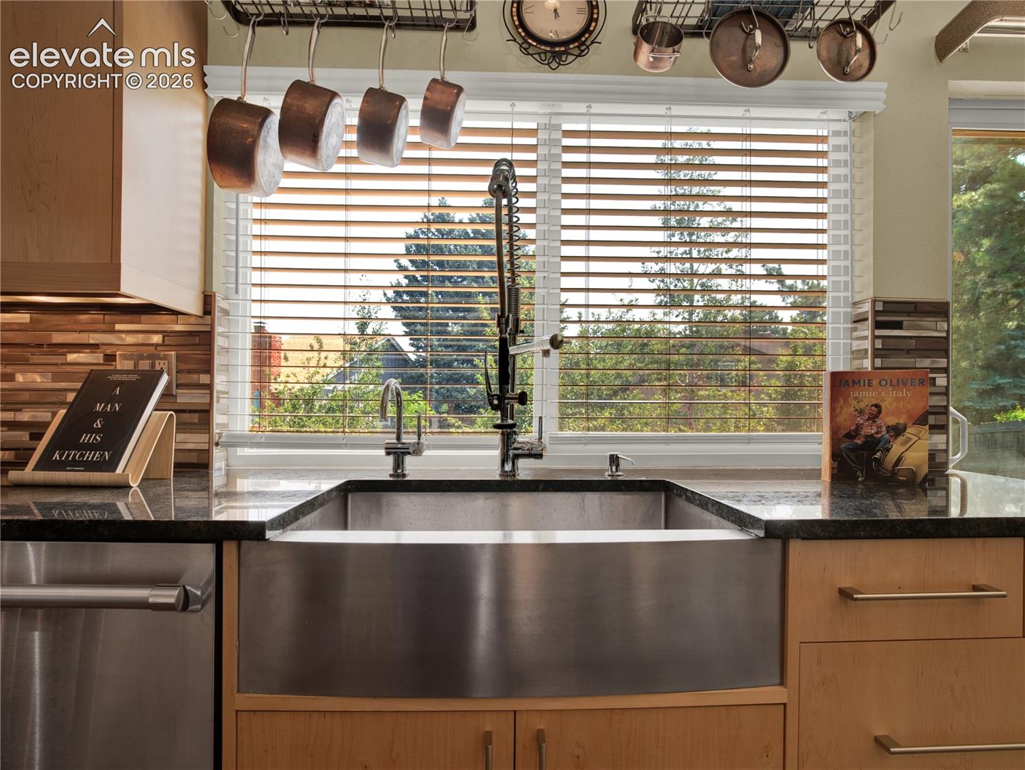 Stainless steel apron-front sink featuring a high-arc pull-down faucet, dark stone countertops, custom light-toned wood cabinetry, and a mosaic tile backsplash