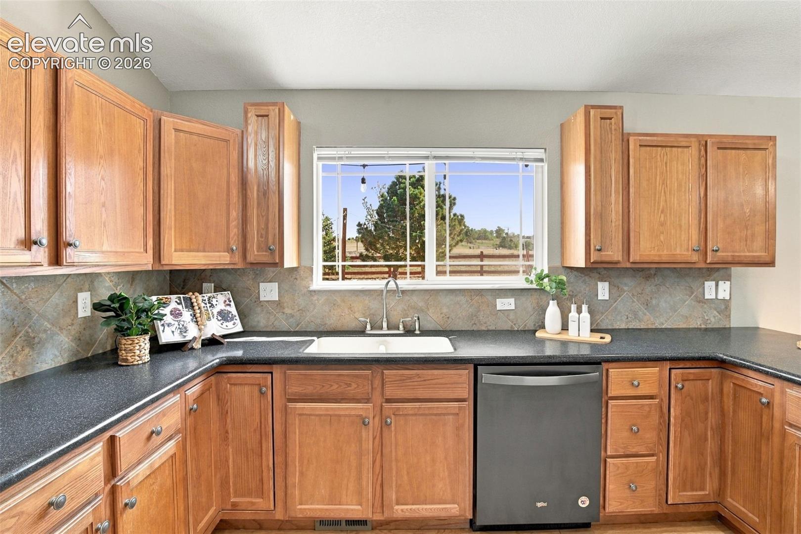 Wood-finish cabinetry with silver-tone hardware, dark countertops, a large window, a tile backsplash, and a built-in dishwasher