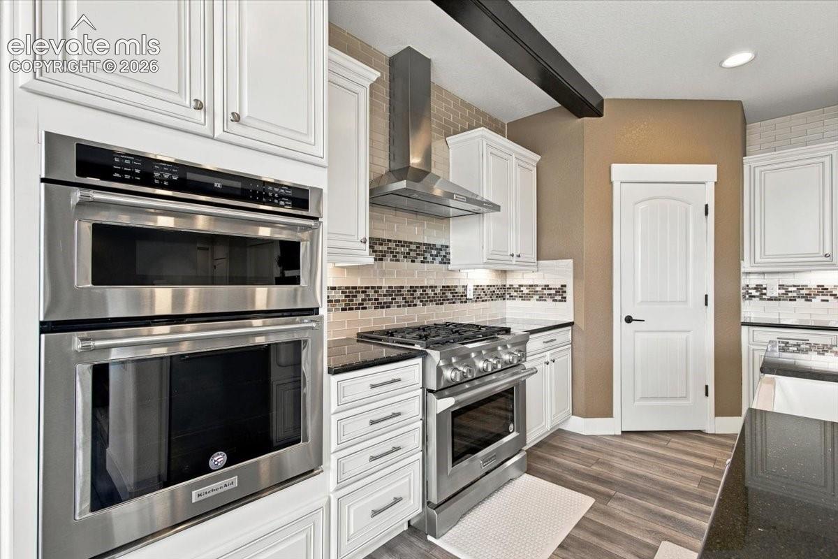 Kitchen featuring white cabinetry, stainless steel double wall ovens, a gas range with a stainless steel vent hood, dark stone countertops, and wood-finish flooring