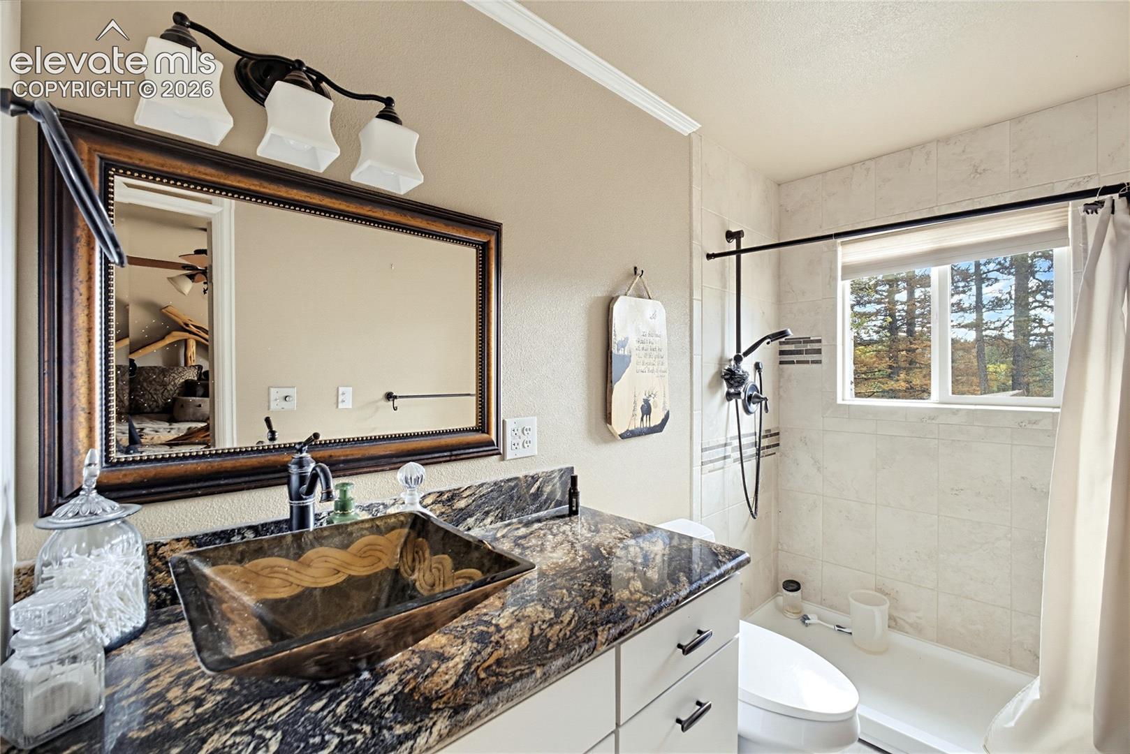 Bathroom featuring a vanity with dark-toned stone-finish countertop, decorative vessel sink, three-light wall-mounted fixture, and a tile-surround shower with sliding window
