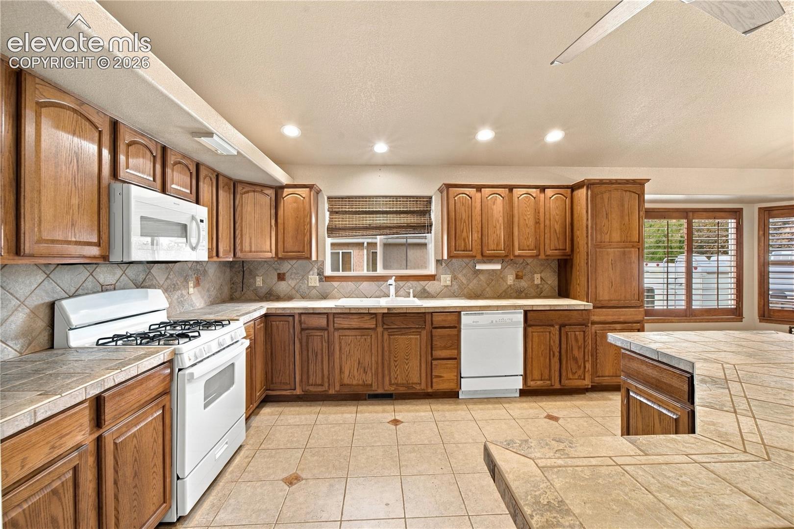 Kitchen featuring wood cabinetry, tile countertops with a matching backsplash, tile flooring with decorative inlay, built-in white appliances, and a ceiling fan