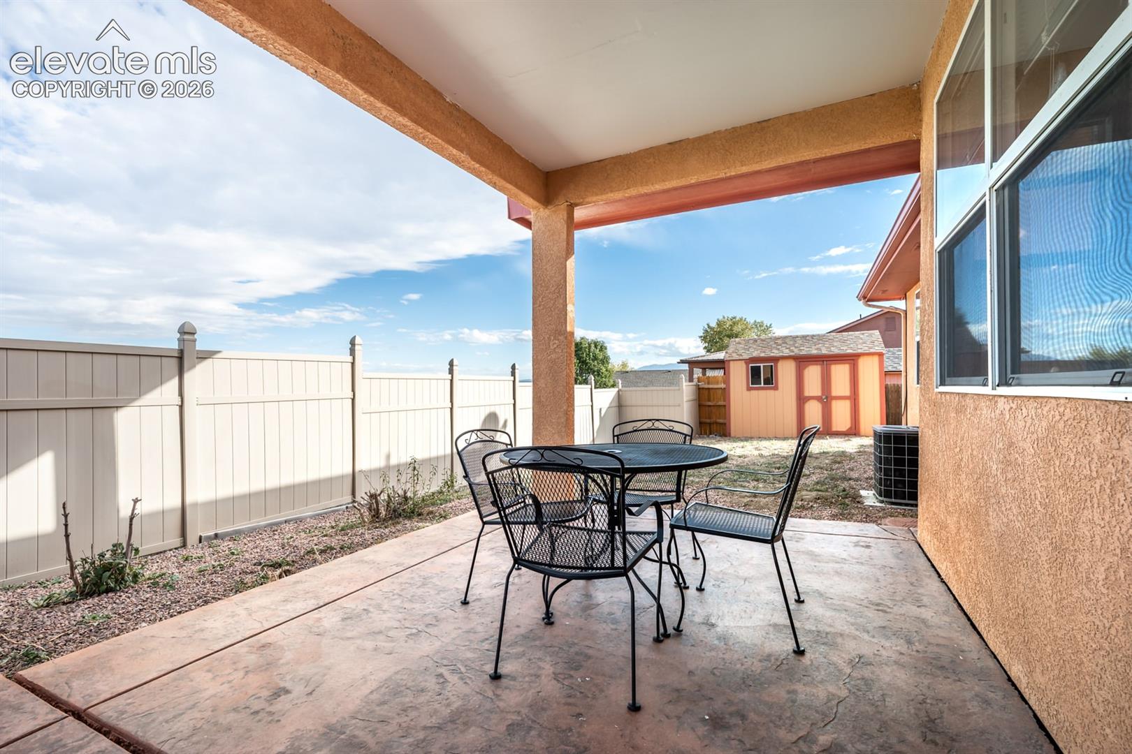 Covered concrete patio featuring a textured finish, enclosed by vinyl fencing with a detached storage shed