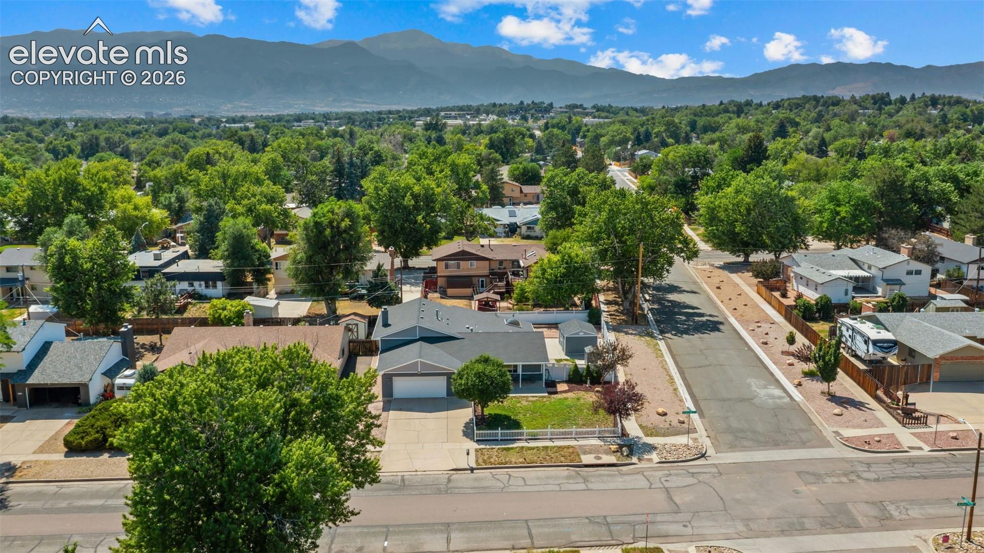Residential street featuring single-family homes, mature trees, mountain backdrop, and clear sky