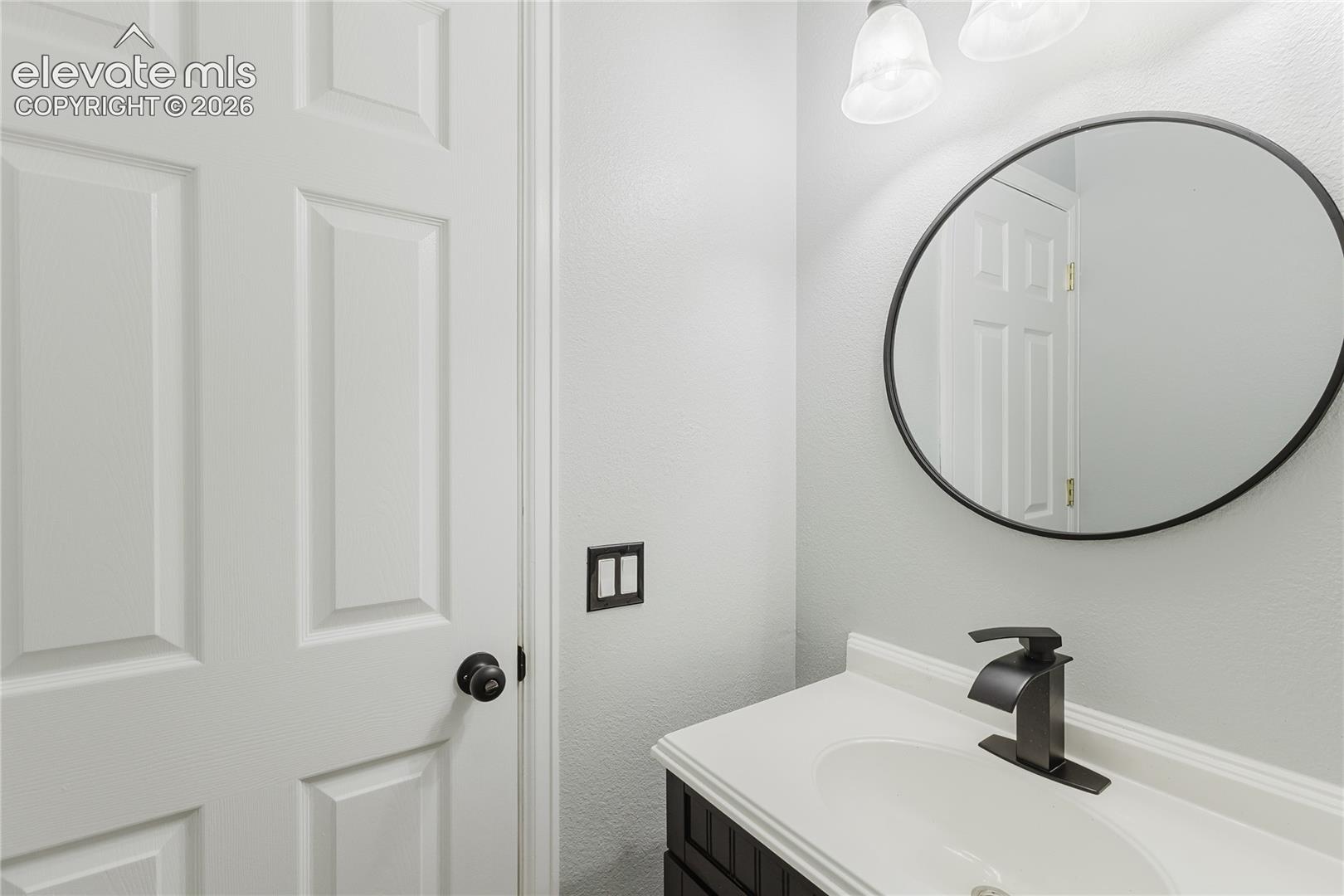 White paneled door with a black doorknob, black framed round mirror, and a black faucet set on a white countertop
