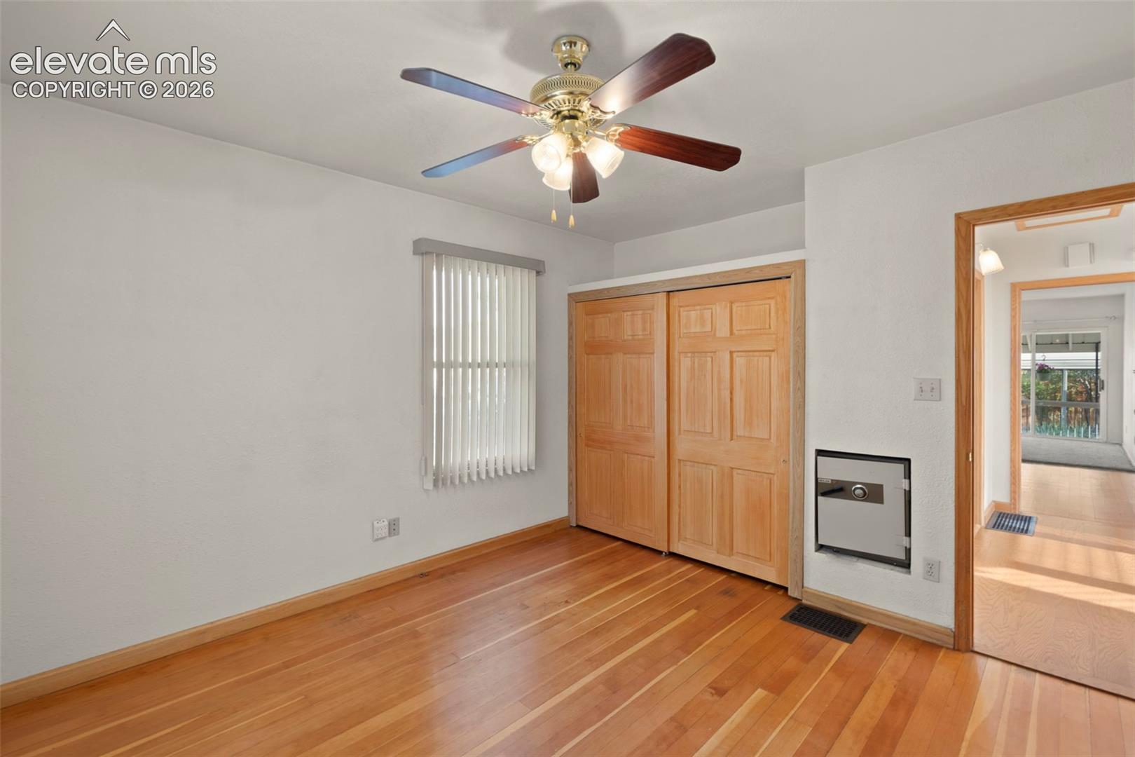 Wood-finish flooring with light-toned baseboards, ceiling fan with light fixture, window with vertical blinds, and a built-in closet with natural wood doors