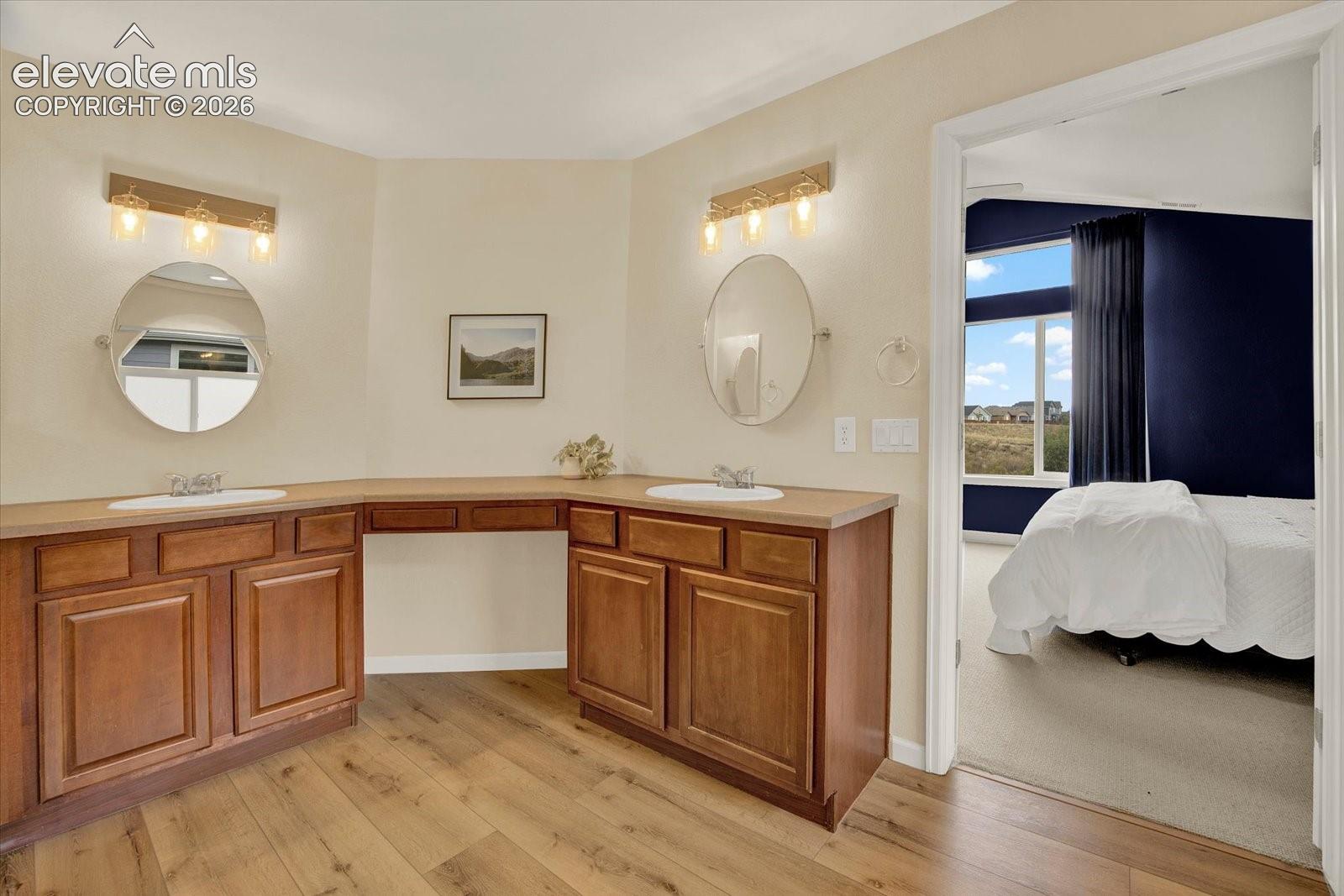 Dual-vanity cabinetry with an integrated makeup desk, solid surface countertops, oval wall-mounted mirrors, and wood-finish flooring