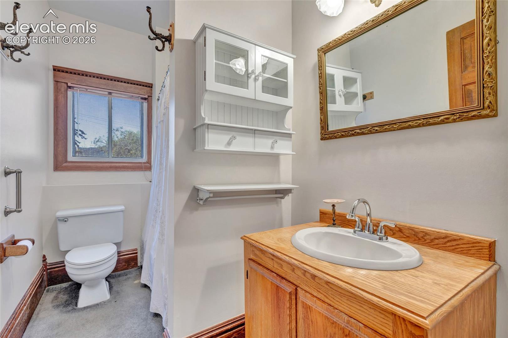 Bathroom featuring a wooden vanity with an integrated basin, wall-mounted white cabinetry with glass-front doors, a wooden-framed mirror, and wood-trimmed window casing