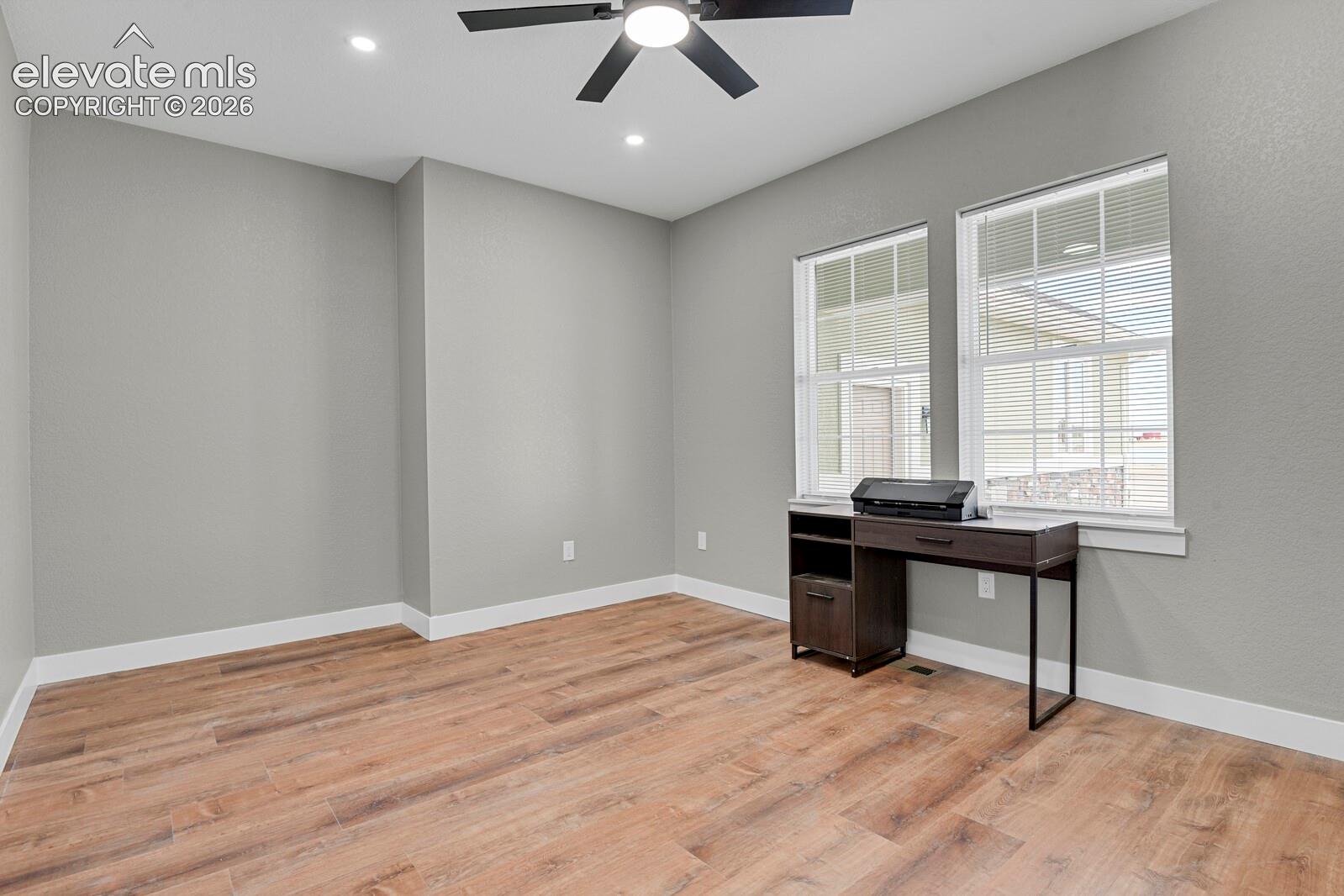 Neutral interior room featuring wood-finish flooring, dual windows with blinds, and a matte black ceiling fan with integrated lighting