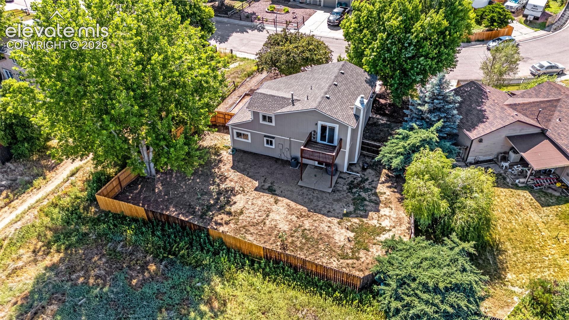 Aerial perspective featuring a multi-level exterior, rear balcony, ground-level concrete patio, fenced perimeter, and mature trees
