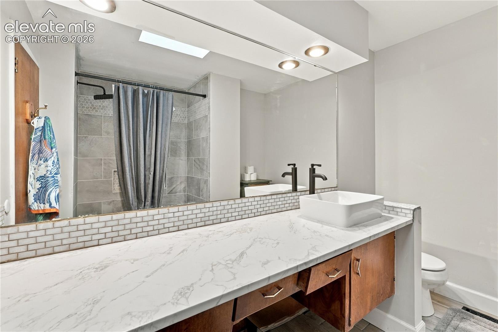 Vanity with a white surface, square vessel sink, and matte black widespread faucet, complemented by a large wall mirror and stone-finish tile shower surround