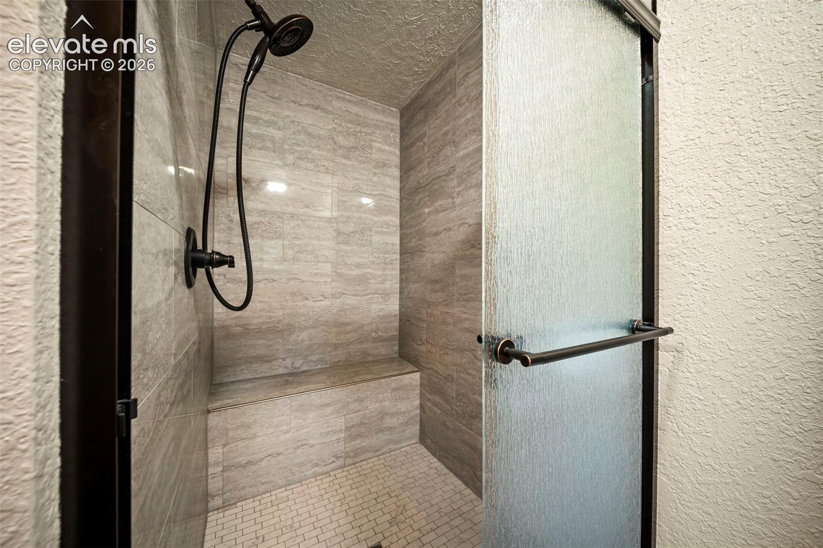 Walk-in shower featuring large-format neutral wall tile, built-in tiled bench, mosaic floor tile, adjustable handheld showerhead, and a textured glass door with a dark metal frame