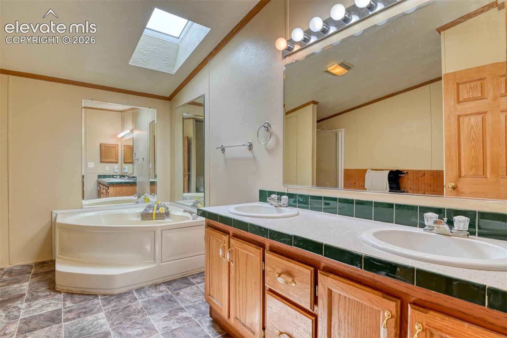 Bathroom featuring a large soaking tub, skylight, dual vanities with integrated sinks, wood-finish cabinetry, and tiled flooring