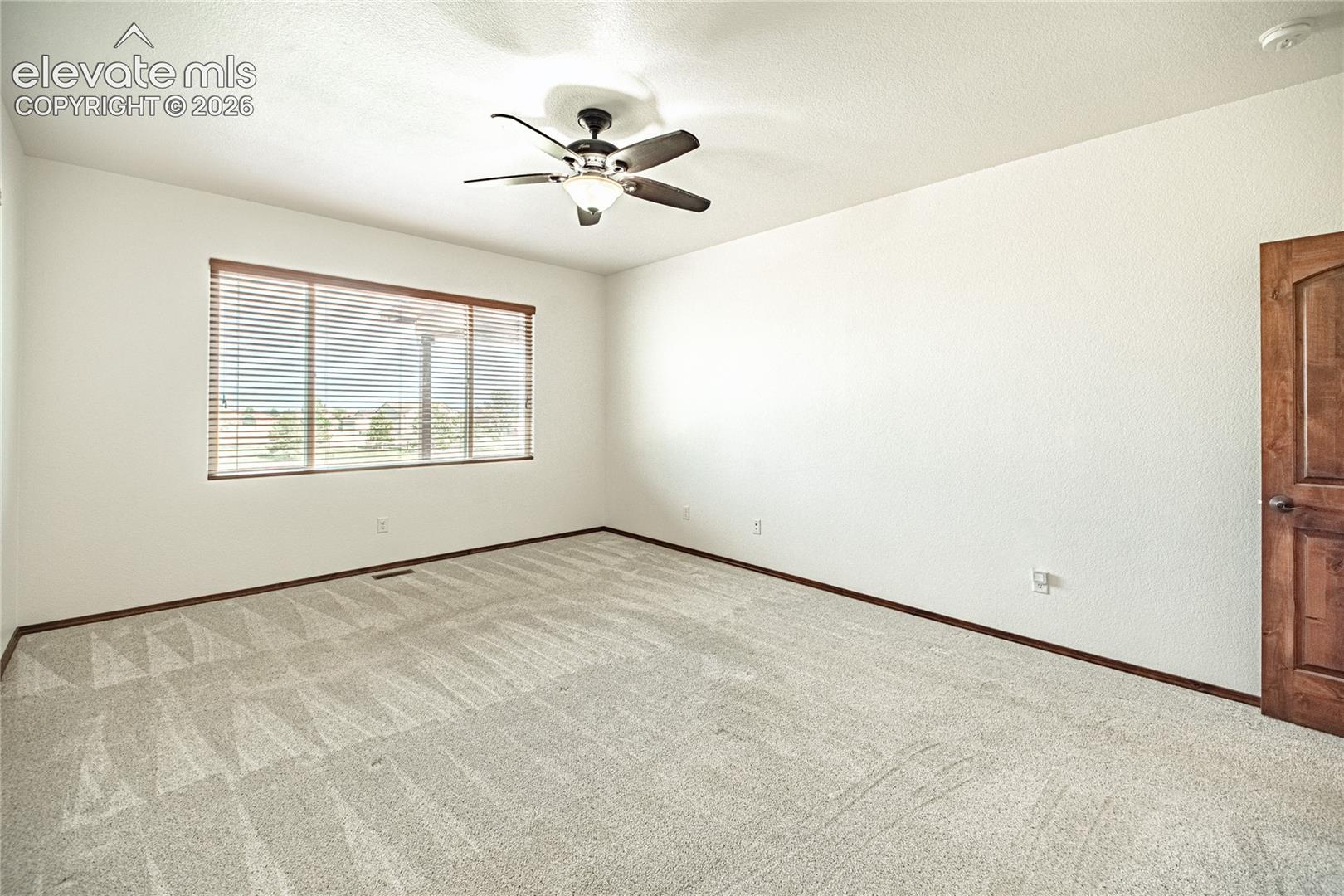 Spacious room featuring light-colored carpet, a large window with horizontal blinds, and a ceiling fan with integrated lighting