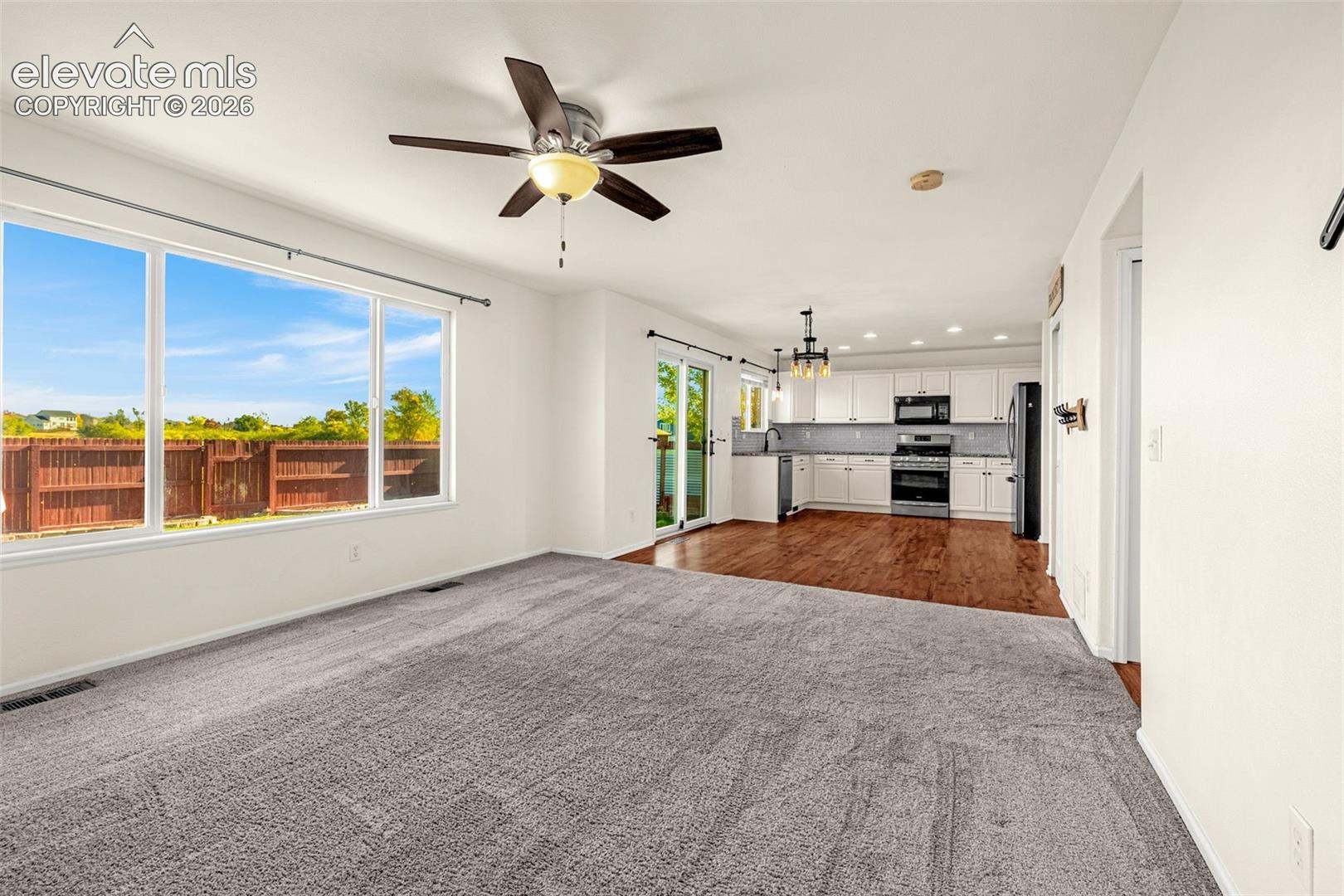 Open-concept space featuring wall-to-wall carpeting, wood-finish flooring in the kitchen, white cabinetry, integrated stainless steel appliances, and a ceiling fan