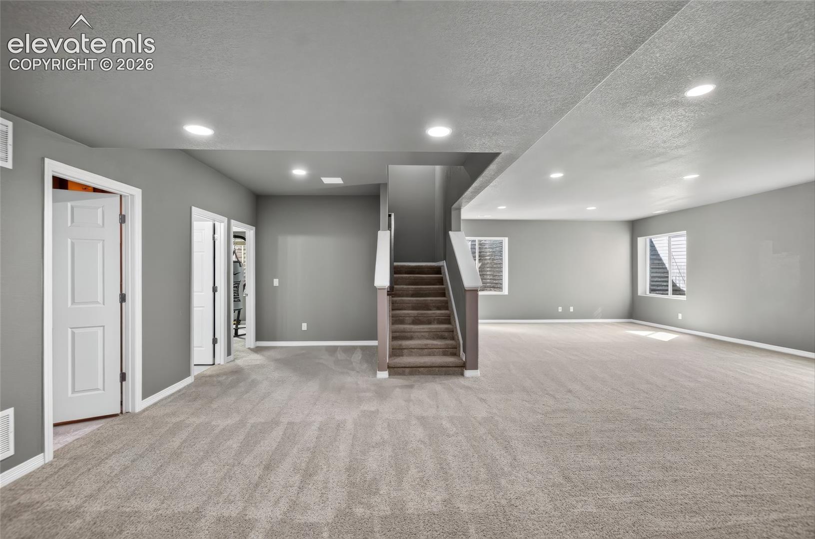 Expansive carpeted area, recessed lighting, two windows with blinds, and a carpeted staircase