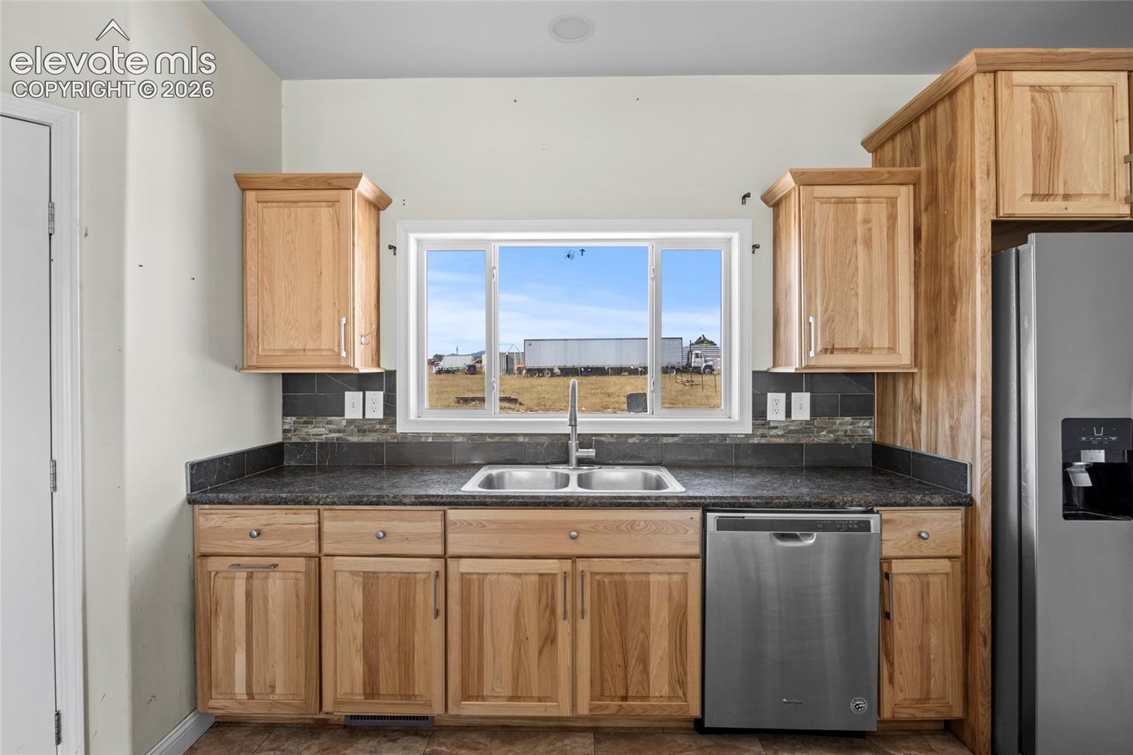 Kitchen featuring natural wood cabinetry, dark stone-finish countertops, stainless steel dishwasher, double-basin sink, and mosaic tile backsplash