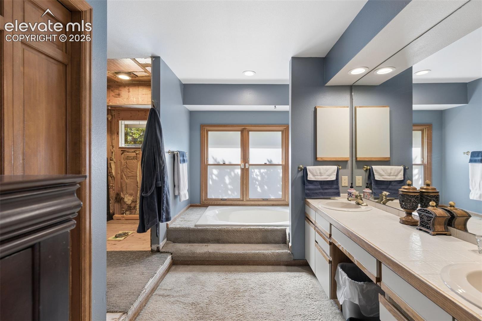 Bathroom featuring a dual-sink vanity with tile countertops, recessed lighting, a full-width vanity mirror, and a soaking tub positioned on a raised platform
