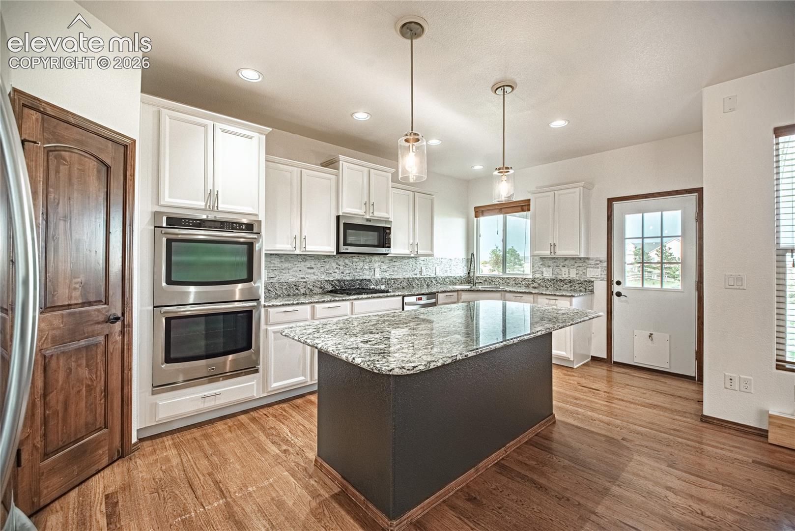 Kitchen featuring a large island with a granite countertop, white cabinetry, stainless steel appliances including double ovens, wood-finish flooring, and pendant lighting