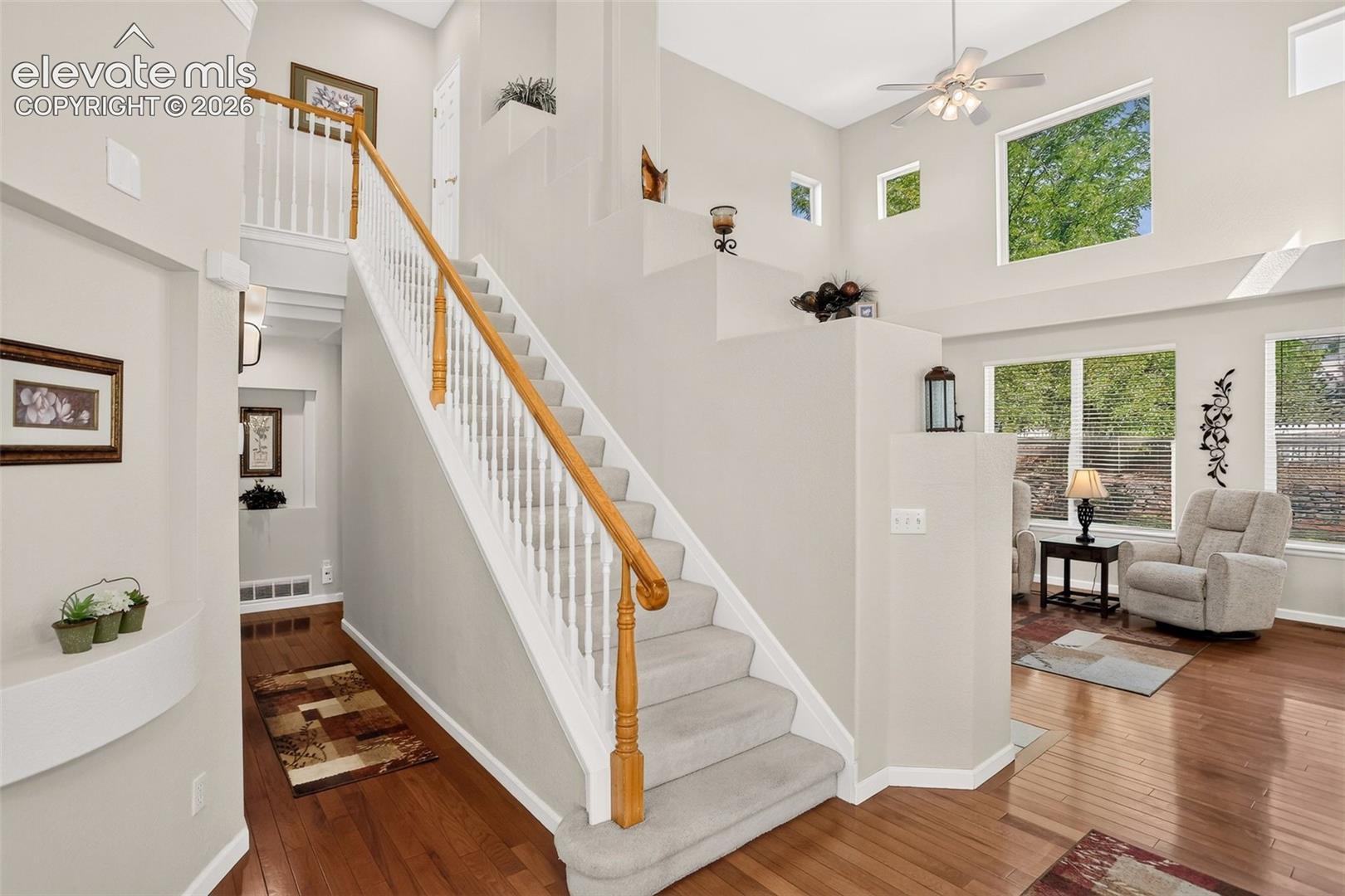 Hardwood-finish flooring, carpeted staircase with white balusters and wood-finish handrail, vaulted ceiling with ceiling fan, and high windows