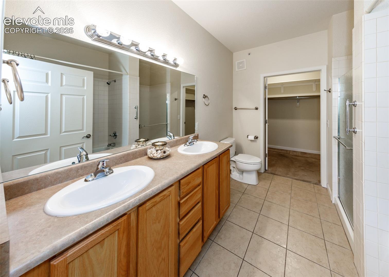 Dual-basin vanity featuring wood cabinetry and solid-surface countertop, full-width mirror, tile flooring, and walk-in closet