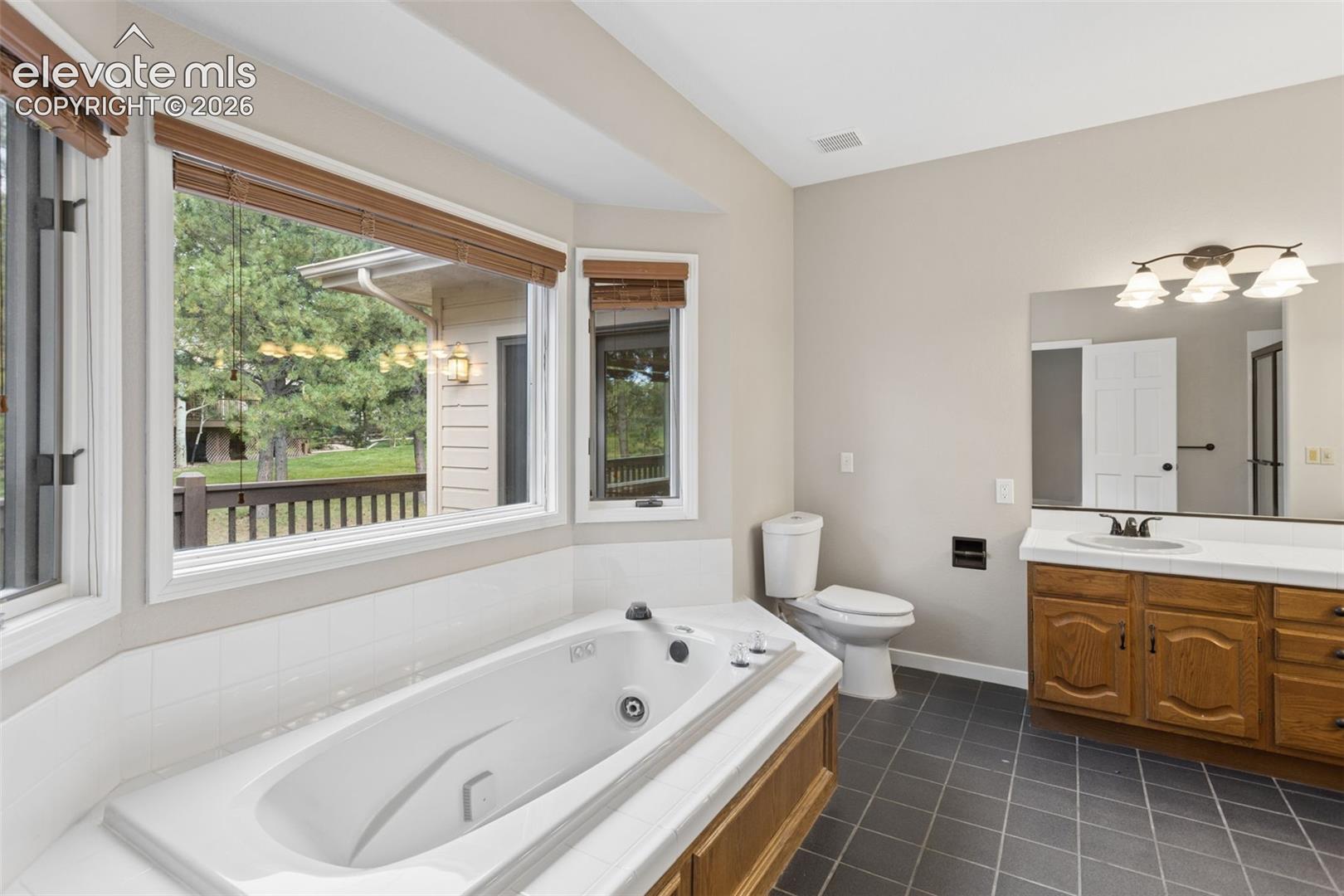 Built-in soaking tub, large vanity with wood-finish cabinetry, integrated single sink, wall-mounted mirror, and tiled flooring