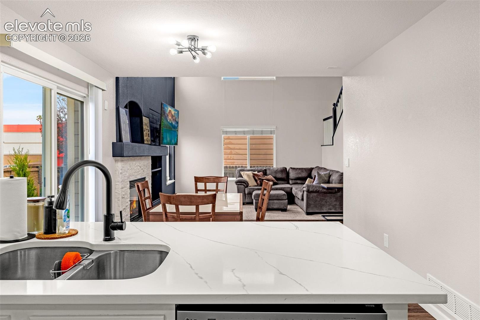 Kitchen featuring a white stone-look countertop, undermount stainless steel double-basin sink, and matte black gooseneck faucet, incorporating a fireplace with a textured surround and a multi-bulb flush-mount light fixture