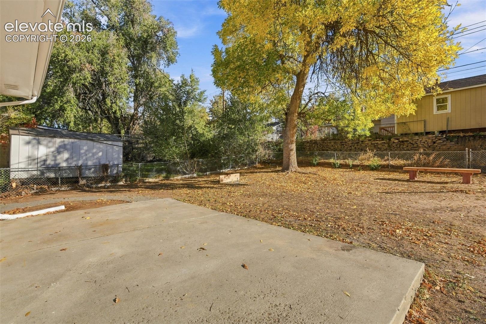 Backyard area featuring a concrete patio, mature trees, chain-link perimeter fencing, and a detached storage structure
