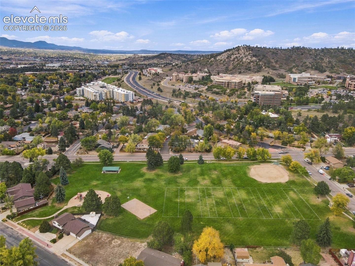Aerial perspective featuring an expansive public park with a manicured athletic field, baseball diamond, playground equipment, and mountain backdrop