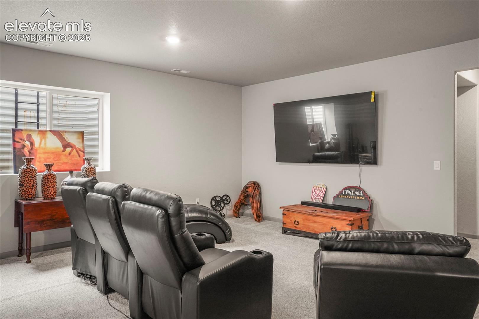 Carpeted entertainment area featuring a wall-mounted display, recessed ceiling lighting, and a window with blinds