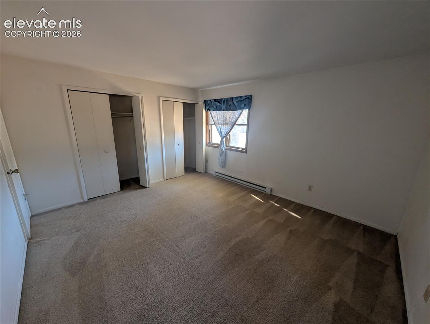 Spacious bedroom featuring neutral carpeting, dual closets with sliding doors, and abundant natural light from corner window with blue valance