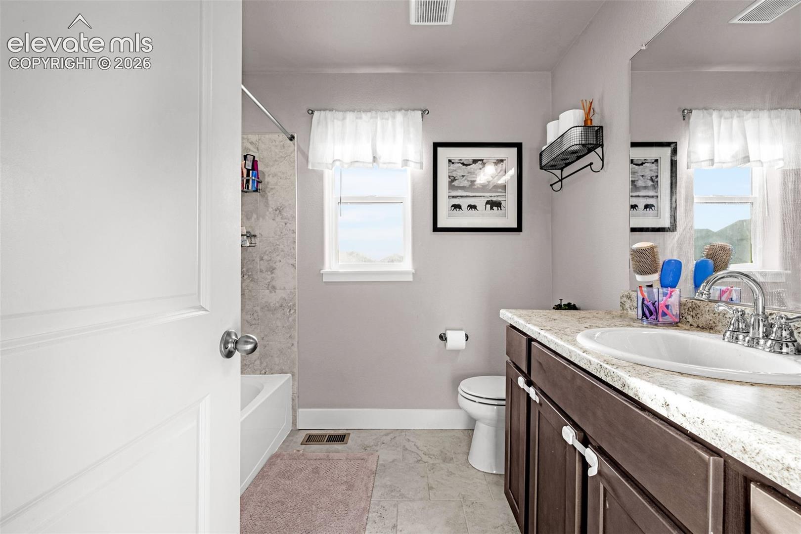 Bathroom featuring a white bathtub with a tiled shower surround, a window with natural light, a dark wood-finish vanity with a light-colored countertop, and a recessed sink with a chrome faucet