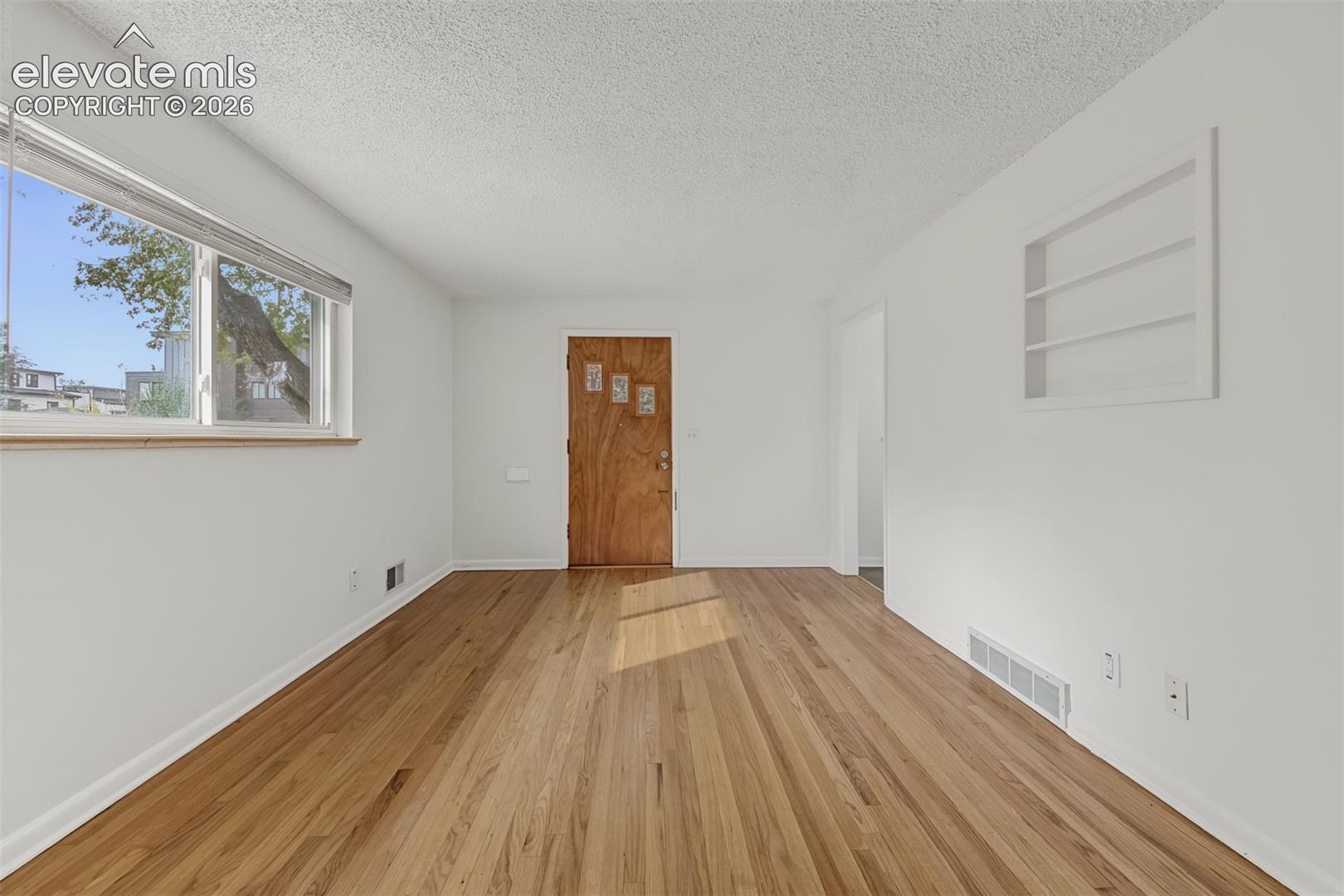 Interior space featuring wood-finish flooring, a wood-grain entry door with decorative glass inserts, large picture window with a natural wood sill, and recessed wall shelving
