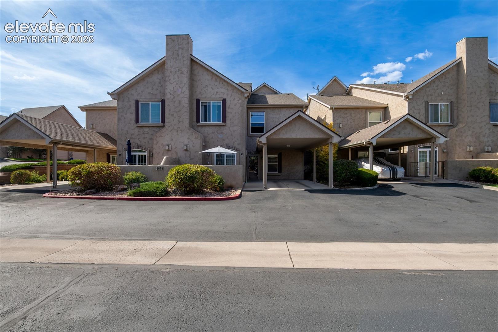 Stucco exterior with multiple covered parking structures, mature landscaping, and an outdoor patio space