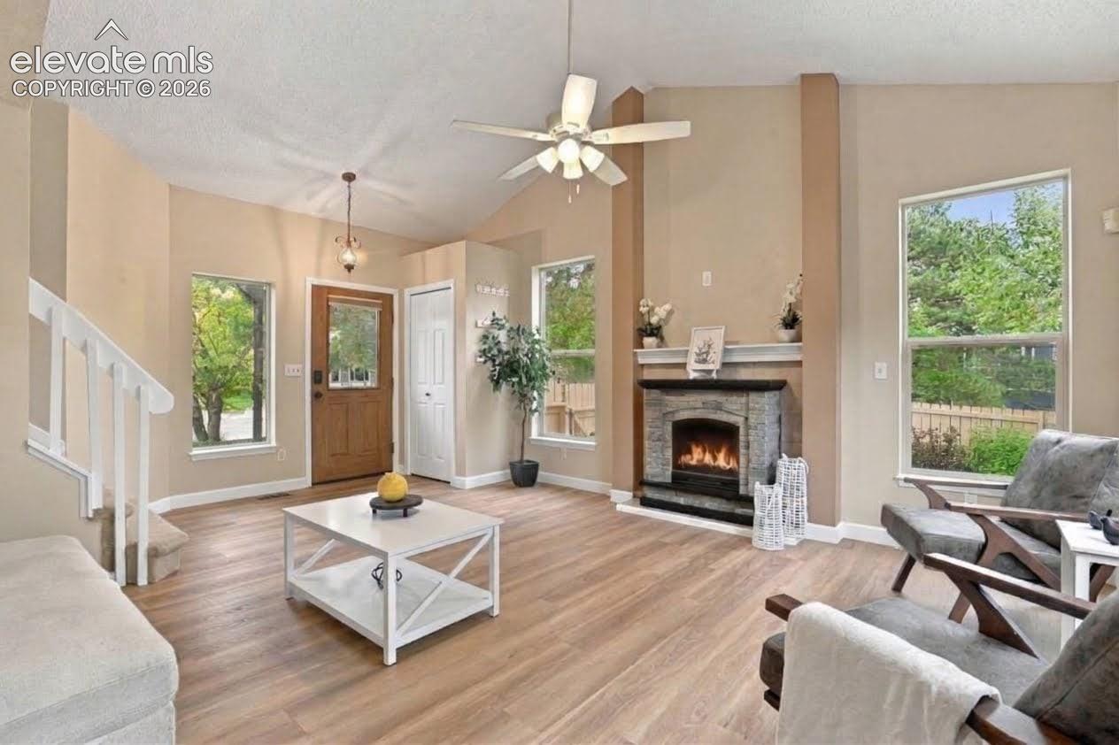 Living area with vaulted ceilings, wood-finish flooring, stone-surround fireplace, ceiling fan, and large windows