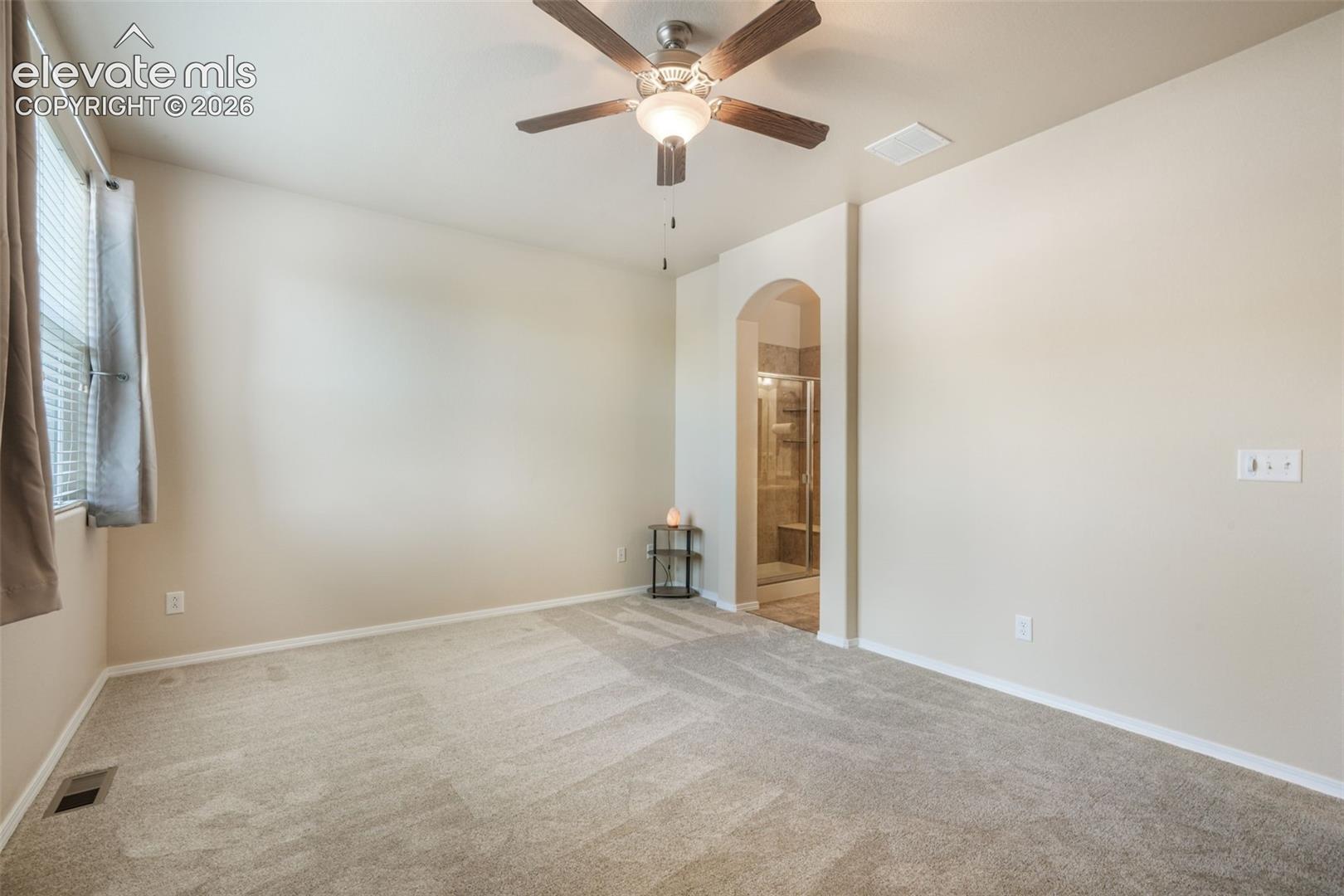 Carpeted room featuring a ceiling fan with integrated lighting, window with blinds, and an arched doorway leading to a bathroom with a glass-enclosed shower
