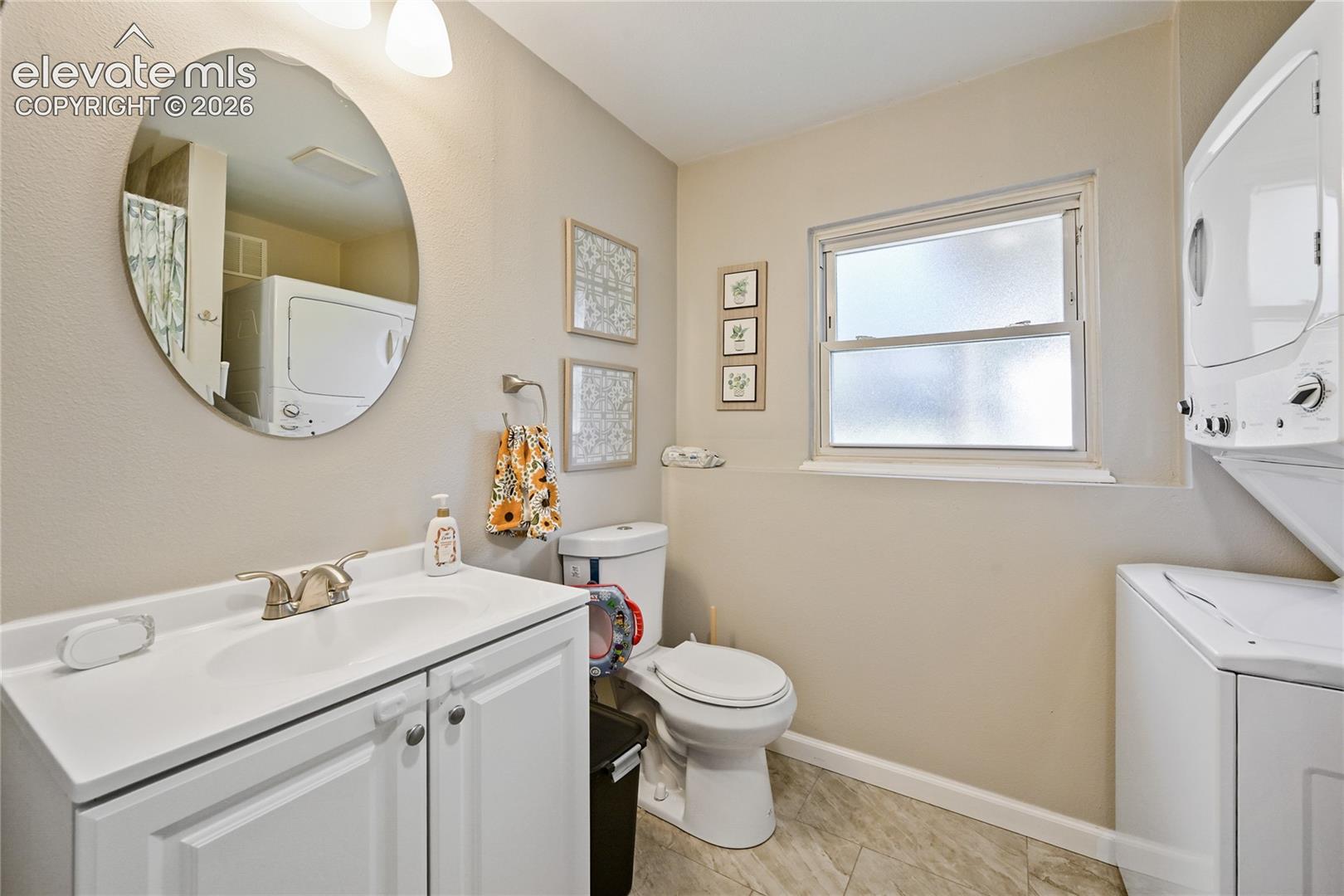 White vanity featuring a cabinet base, integrated basin, and metal faucet, oval wall-mounted mirror, privacy-glass window, and stacked laundry unit