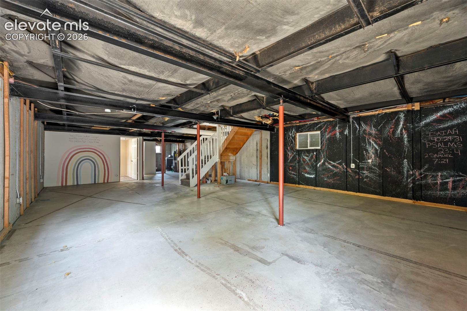 Unfinished lower level featuring exposed steel ceiling beams, concrete flooring, structural support columns, and wooden staircase