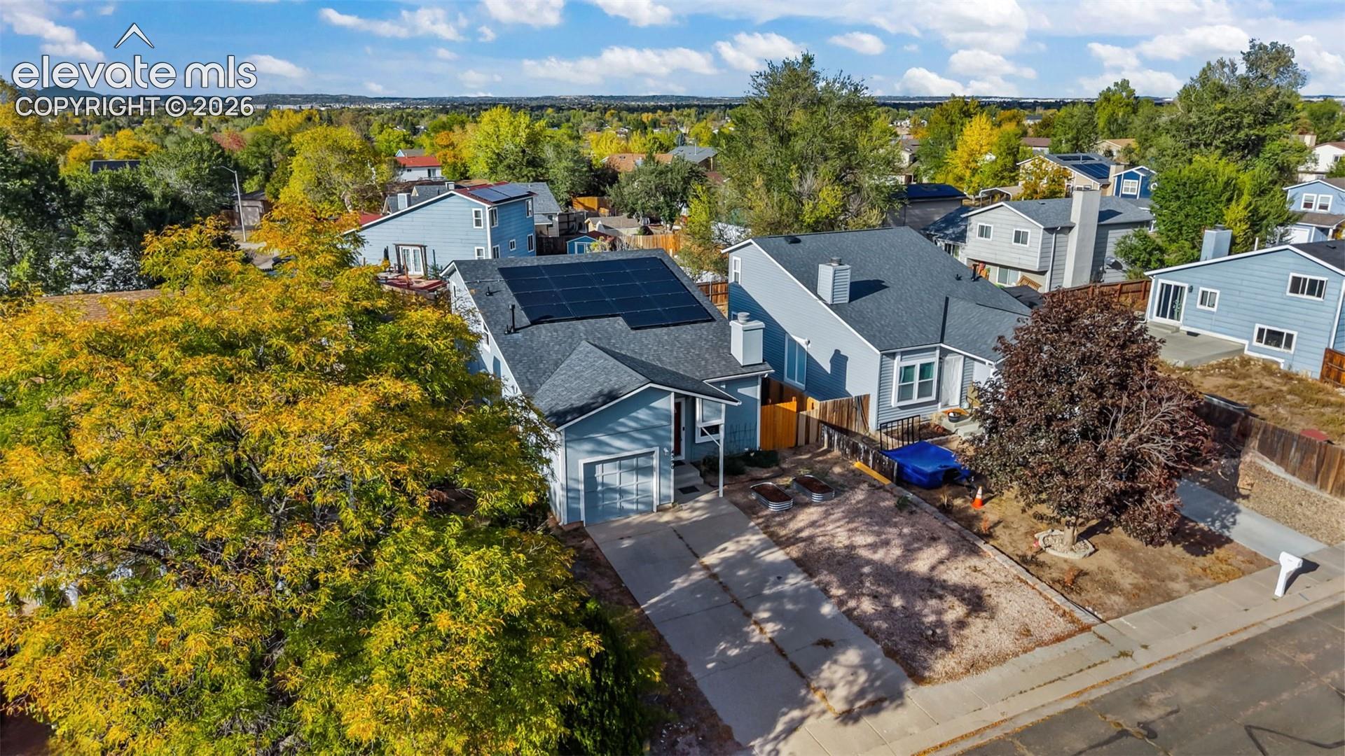 Aerial perspective of single-story residence featuring an asphalt shingle roof, roof-mounted solar panel array, attached garage, and mature tree canopy