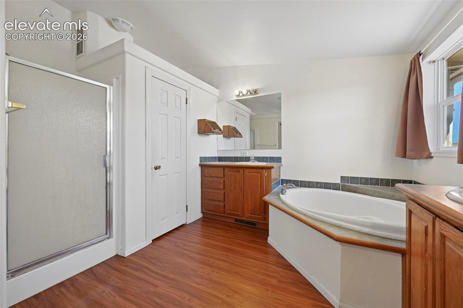 Bathroom featuring wood-finish flooring, a separate walk-in shower with a framed glass door, a built-in jetted soaking tub with tile surround, and wood cabinetry with a countertop basin