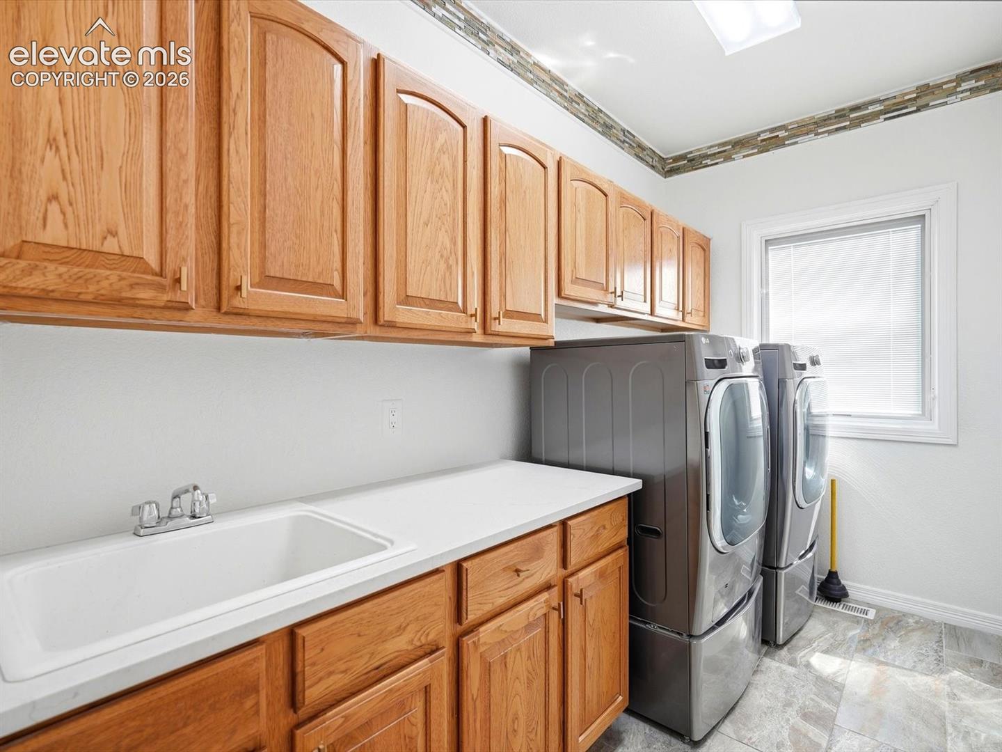 Laundry space featuring wood-finish cabinetry, solid-surface countertop, integrated utility sink with chrome faucet, and tile flooring