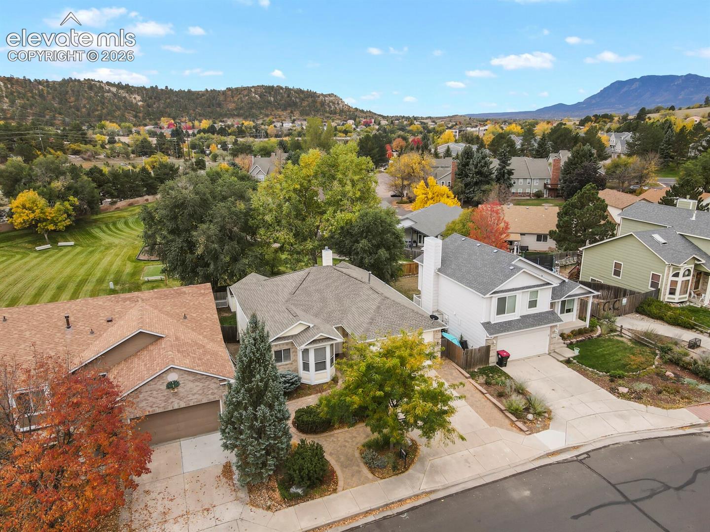 Aerial perspective featuring single-family residences with pitched shingle roofing, concrete driveways, manicured landscaping, and mature trees against a mountainous backdrop
