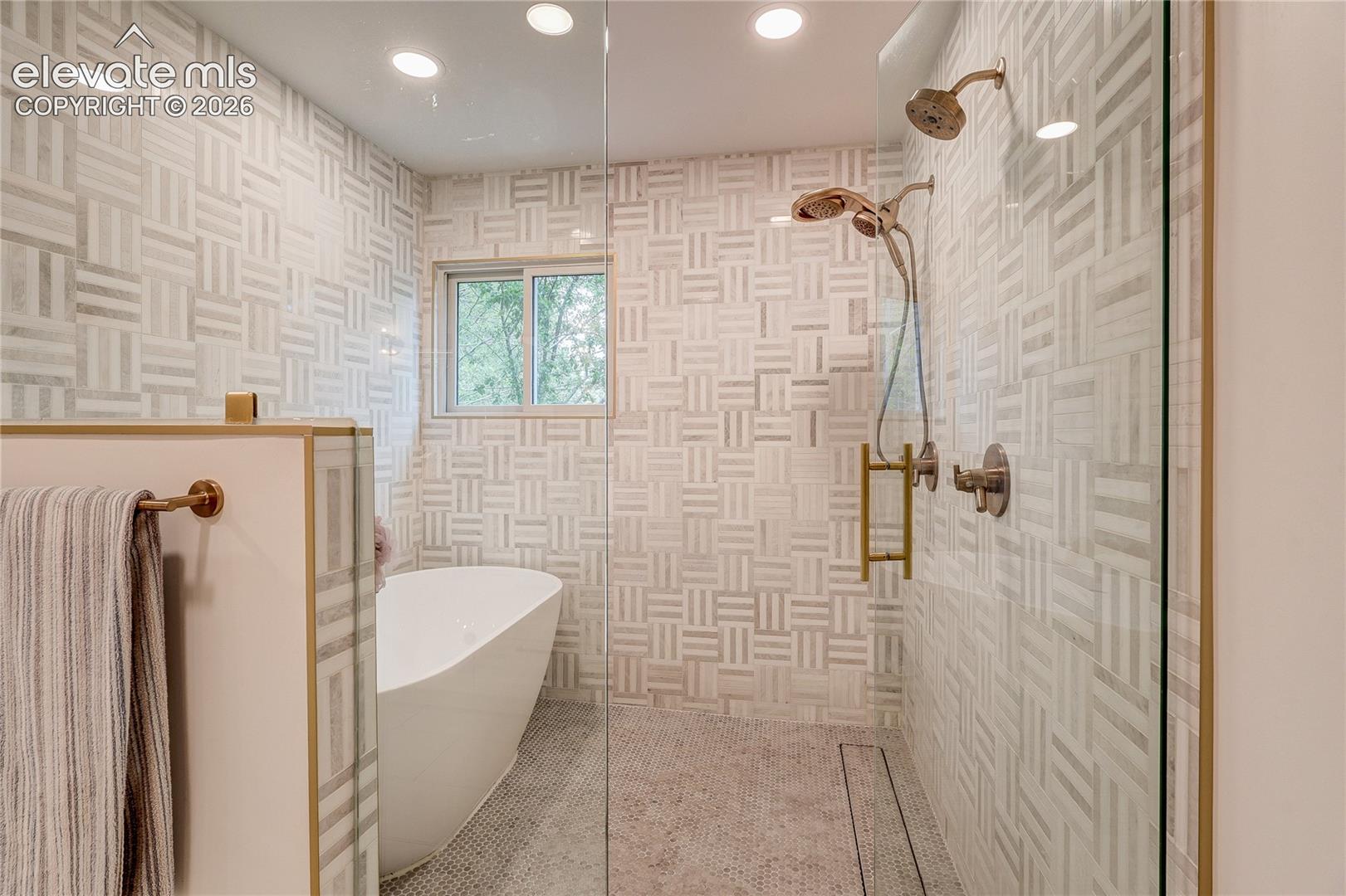 Floor-to-ceiling geometric-patterned wall tile, walk-in shower with glass enclosure, dual shower heads, freestanding soaking tub, and natural light from a rectangular window
