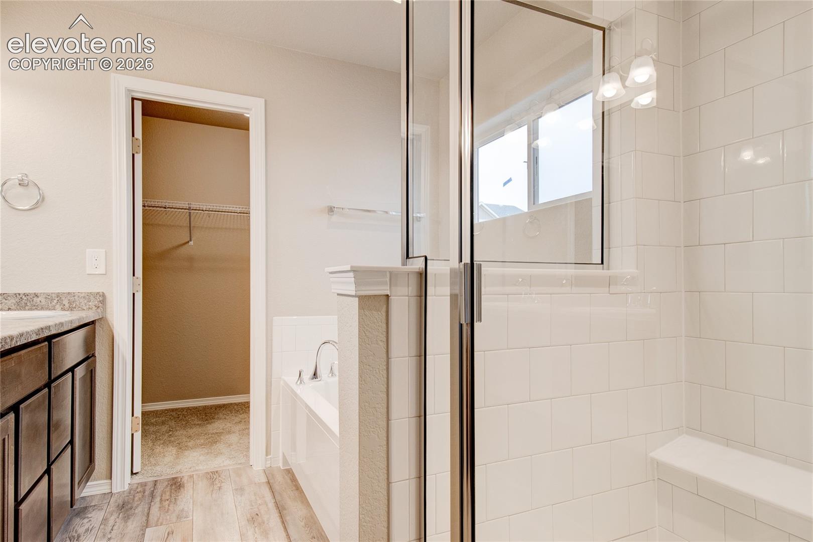 Bathroom featuring a glass-enclosed shower with white subway tile, integrated bench seating, wood-finish flooring, a neutral-toned countertop with an under-mount sink, and dark wood cabinetry