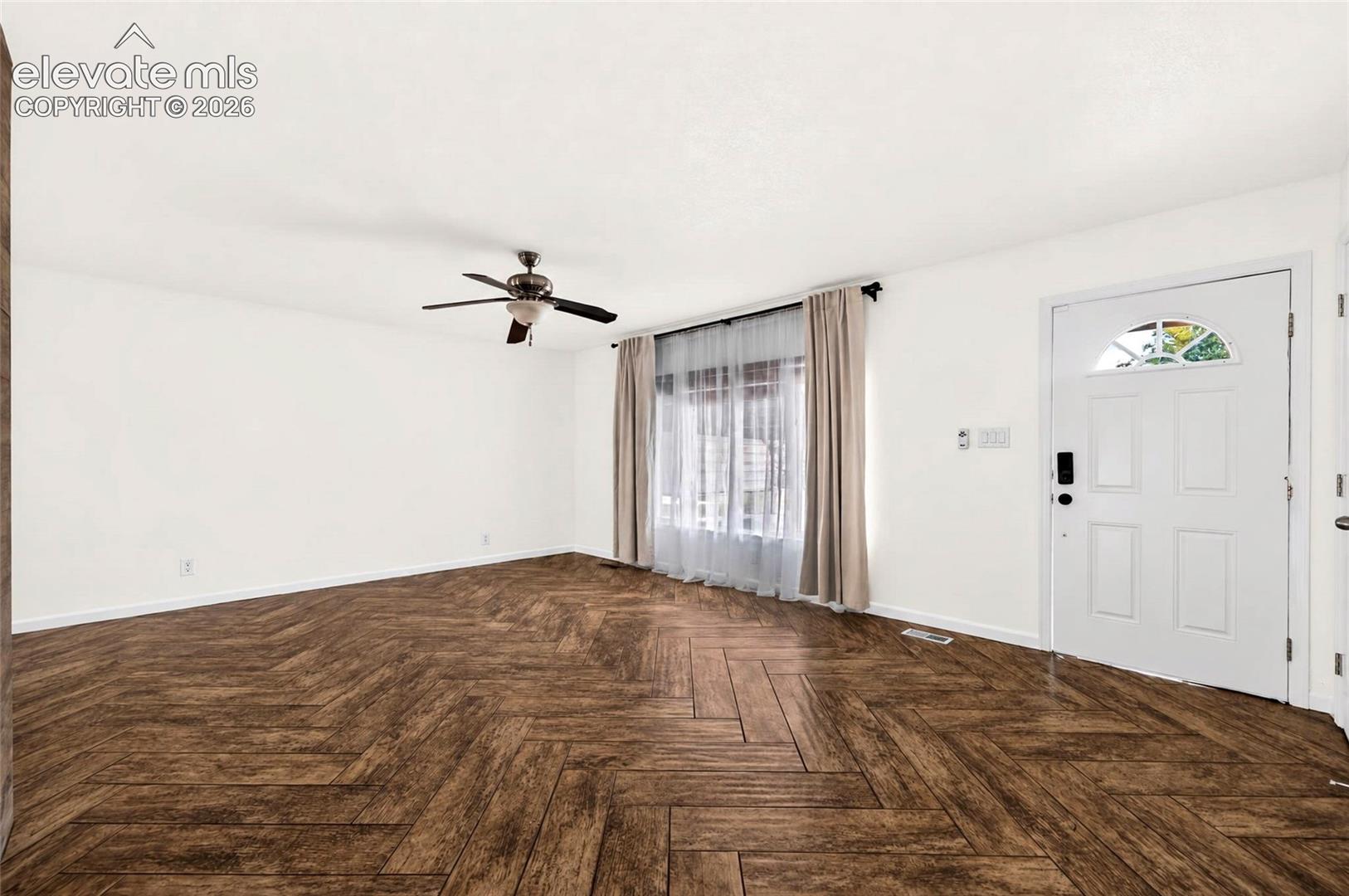 Herringbone wood-finish flooring, white walls, ceiling fan with integrated lighting, and a solid-core entry door featuring a semi-circular transom window