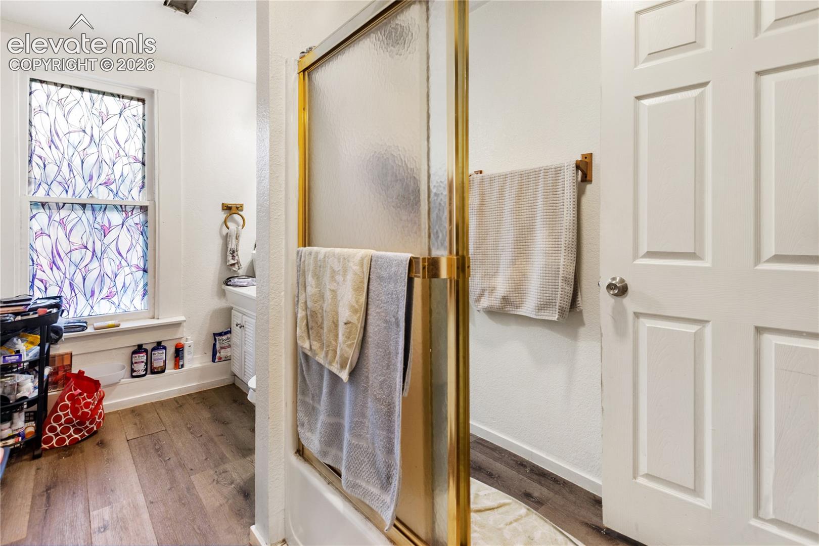 Bathroom featuring wood-finish flooring, a shower with a gold-tone frame and textured glass enclosure, a six-panel door, and a window with patterned privacy film
