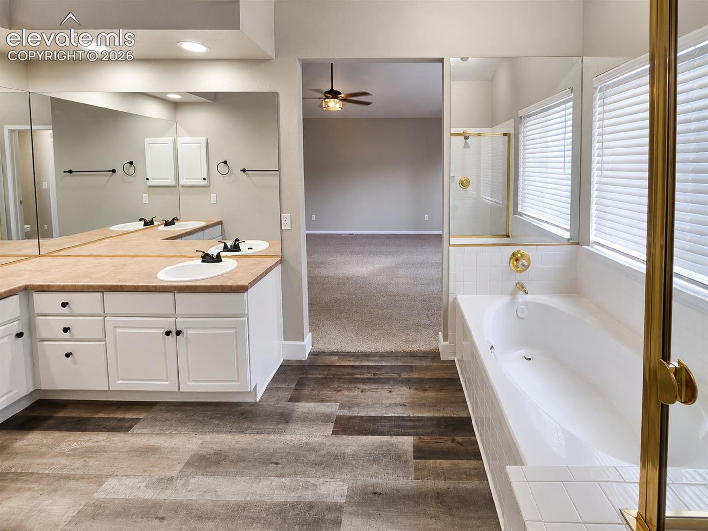 Soaking tub with white tile surround, adjacent shower with gold-finish frame, and wood-finish flooring
